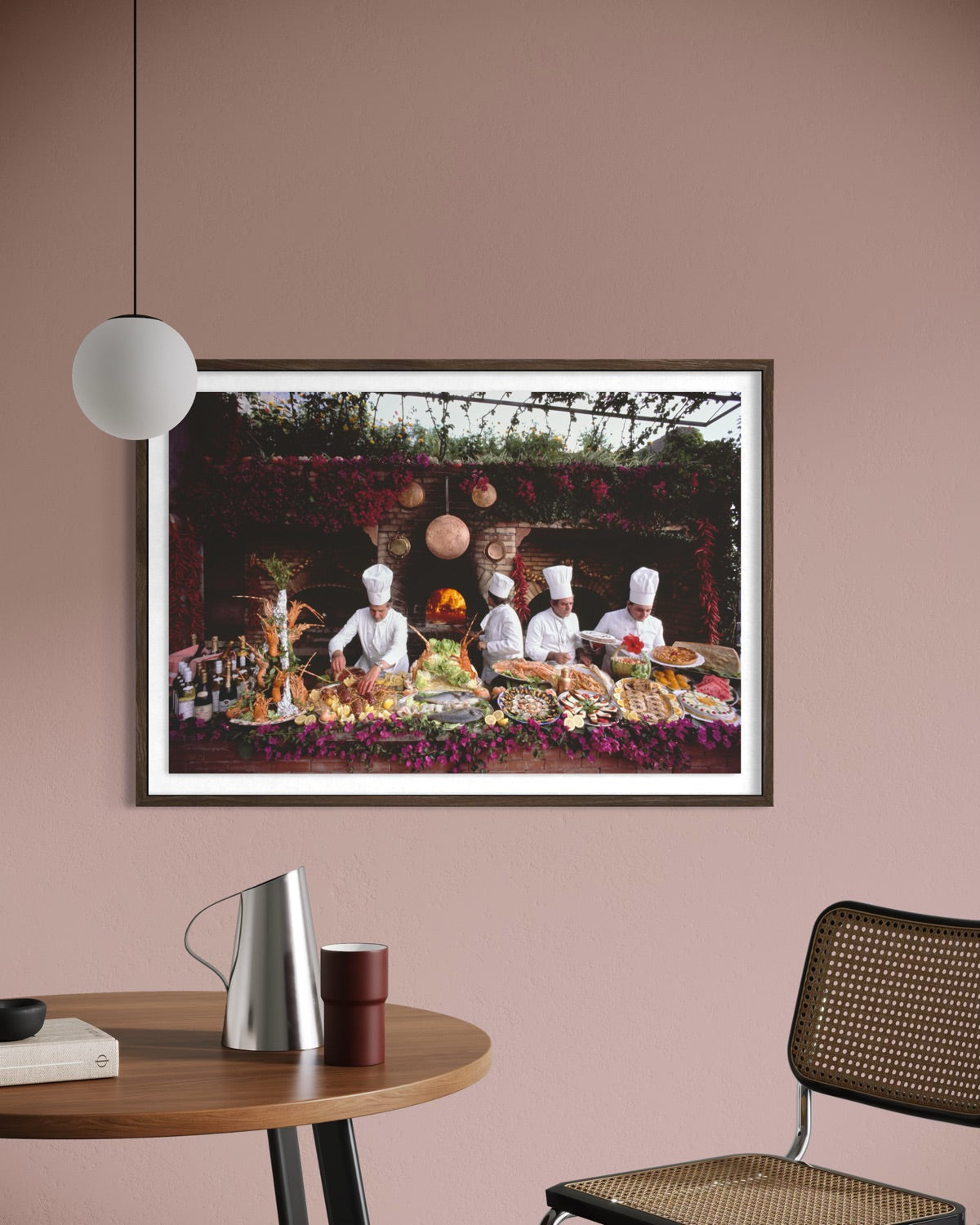 A framed art print depicting chefs in white uniforms serving a large buffet table, displayed on a dusty pink wall above a wooden table with a silver pitcher and a woven chair. The Picturalist Wall Art and Photography for Designed Interiors. Getty Images Gallery.