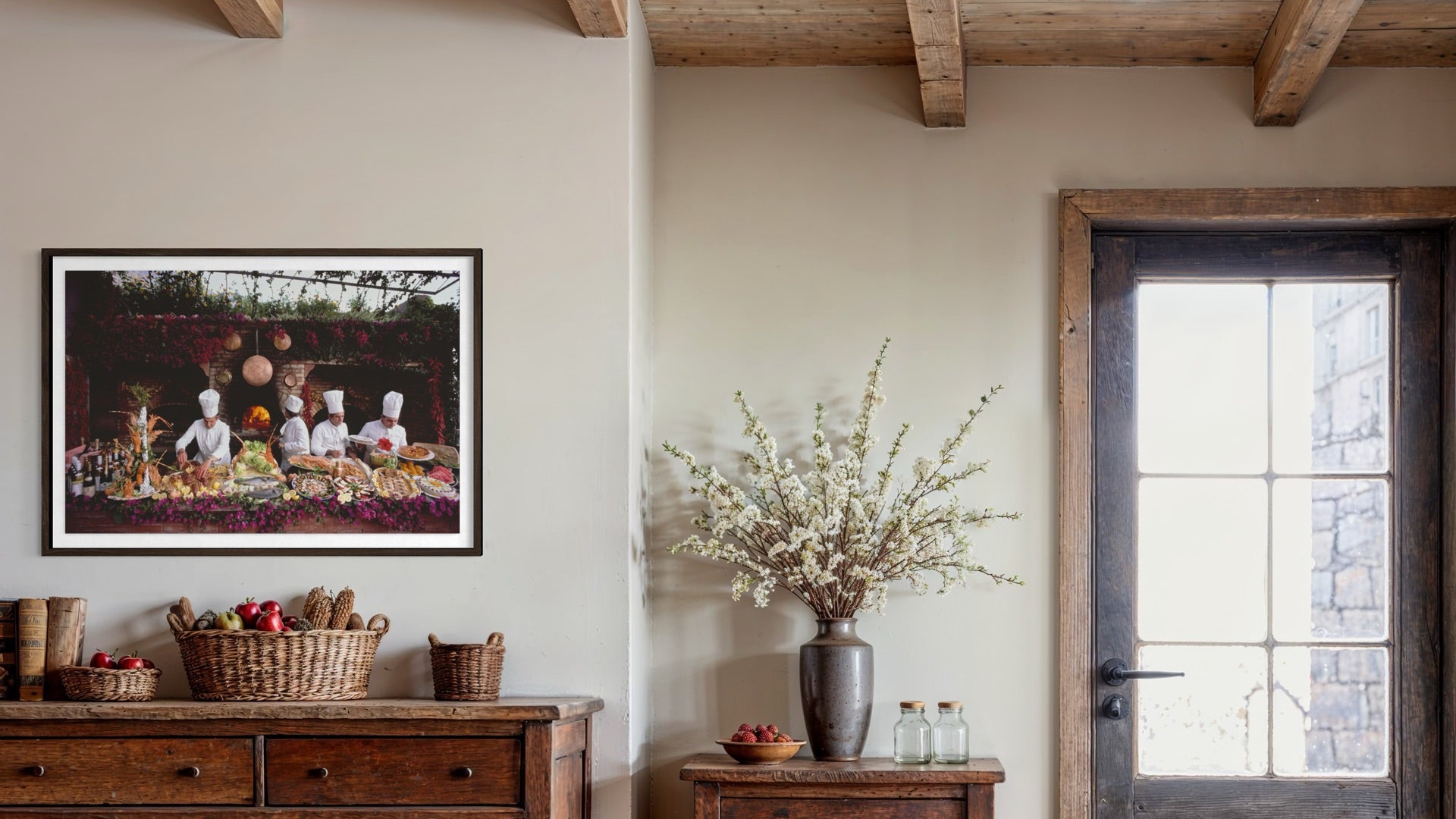 A framed art print depicting a rustic outdoor picnic scene with figures and food, displayed on a beige wall in a styled room with a wooden dresser, a vase of white blossoms, and a dark wood door. The Picturalist Wall Art and Photography for Designed Interiors. Getty Images Gallery.