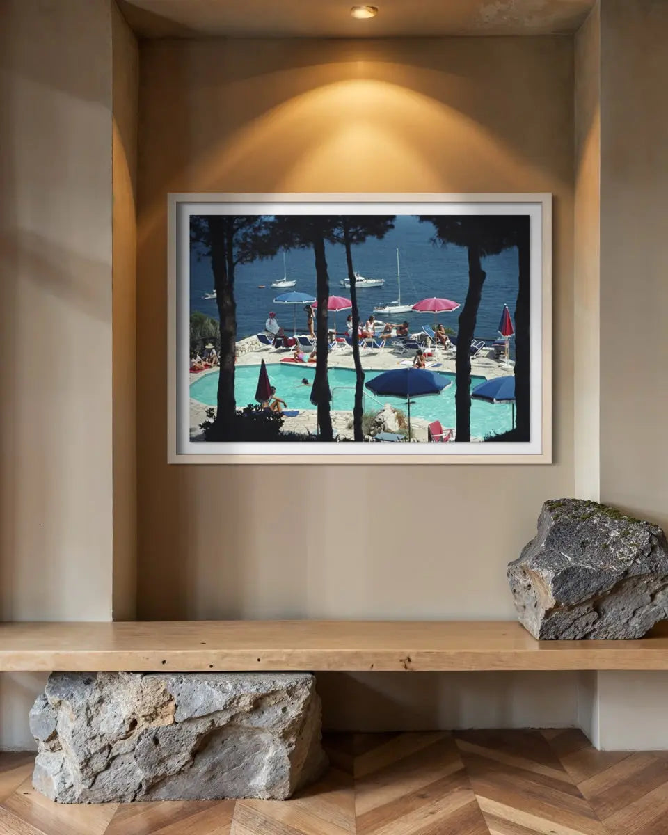 A framed art print depicting a sunny poolside scene with blue water, umbrellas, and tall dark trees, displayed on a beige wall above a wooden bench with decorative rocks.