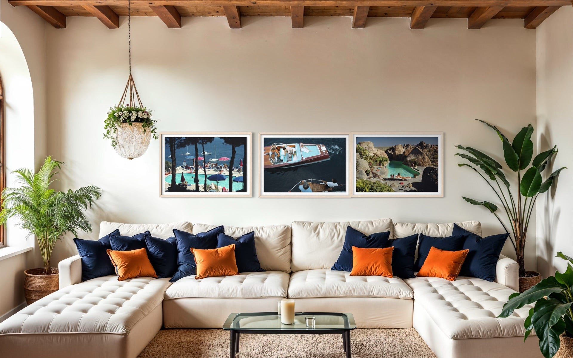 A styled living room featuring a cream tufted sectional sofa with navy and orange accent pillows, a glass coffee table, and three framed landscape photographs on the wall above the couch.