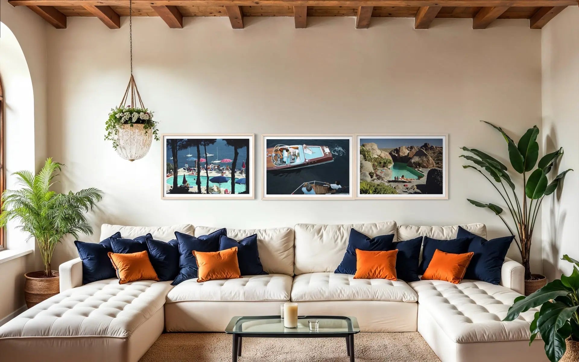 A styled living room featuring a cream tufted sectional sofa with navy and orange accent pillows, a glass coffee table, and three framed landscape photographs on the wall above the couch.