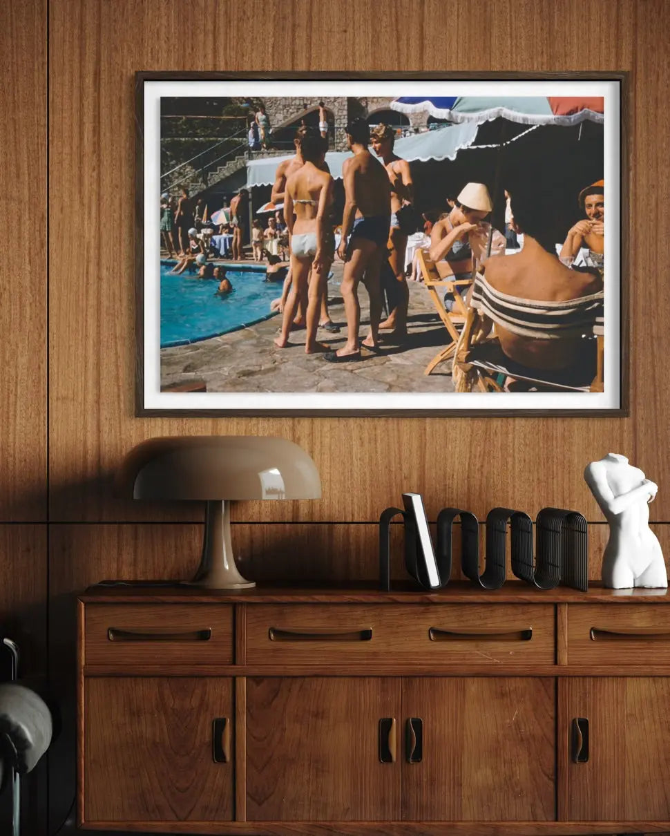 A framed photographic print of a vintage poolside scene with people, displayed on a wood-paneled wall above a wooden console table with a lamp and decorative objects.