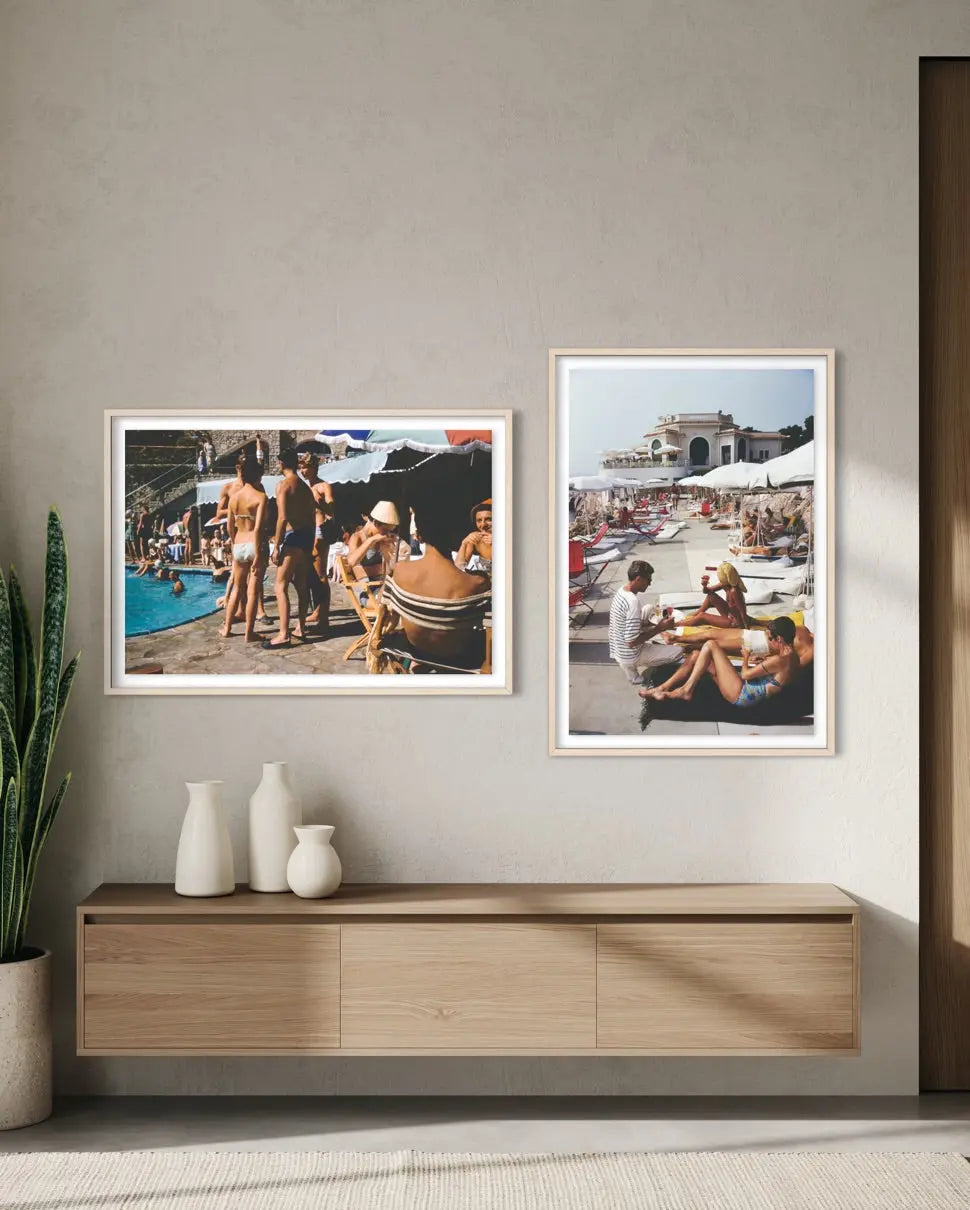 Two framed vintage beach and poolside photographs displayed side-by-side on a textured beige wall above a light wood console table with white ceramic vases.