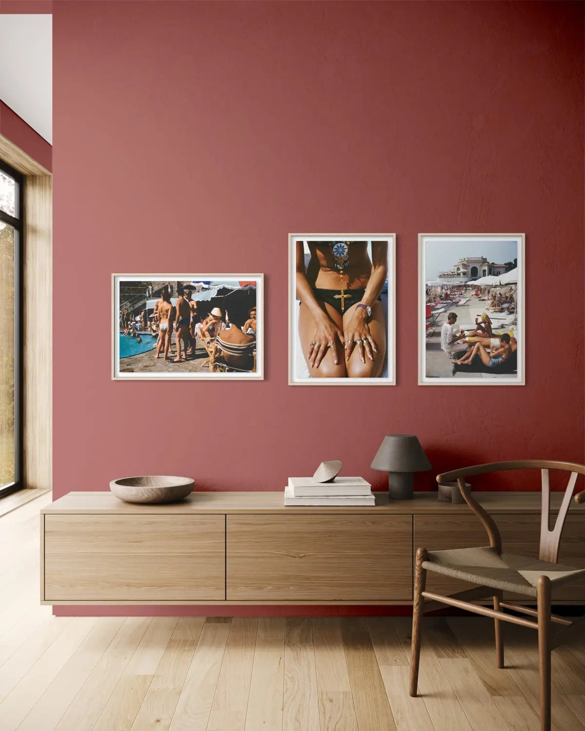 Three framed art prints with white frames are displayed on a textured terracotta red wall above a light wood console table. The room features light wood floors, a wooden chair, and a small table lamp.