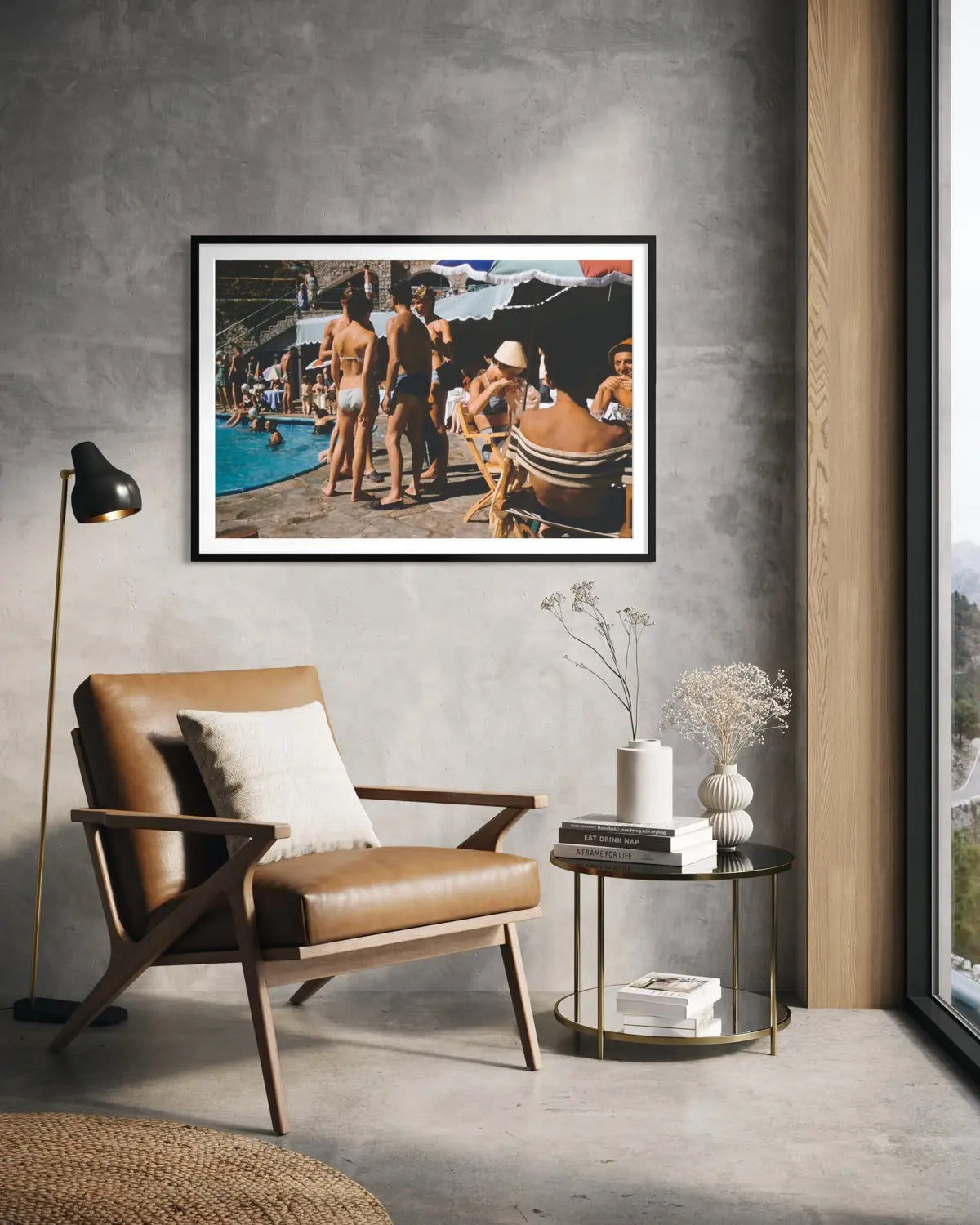 A framed art print depicting a vintage beach scene with people by a pool, displayed on a textured gray wall in a modern living room setting with a brown leather armchair and a side table.