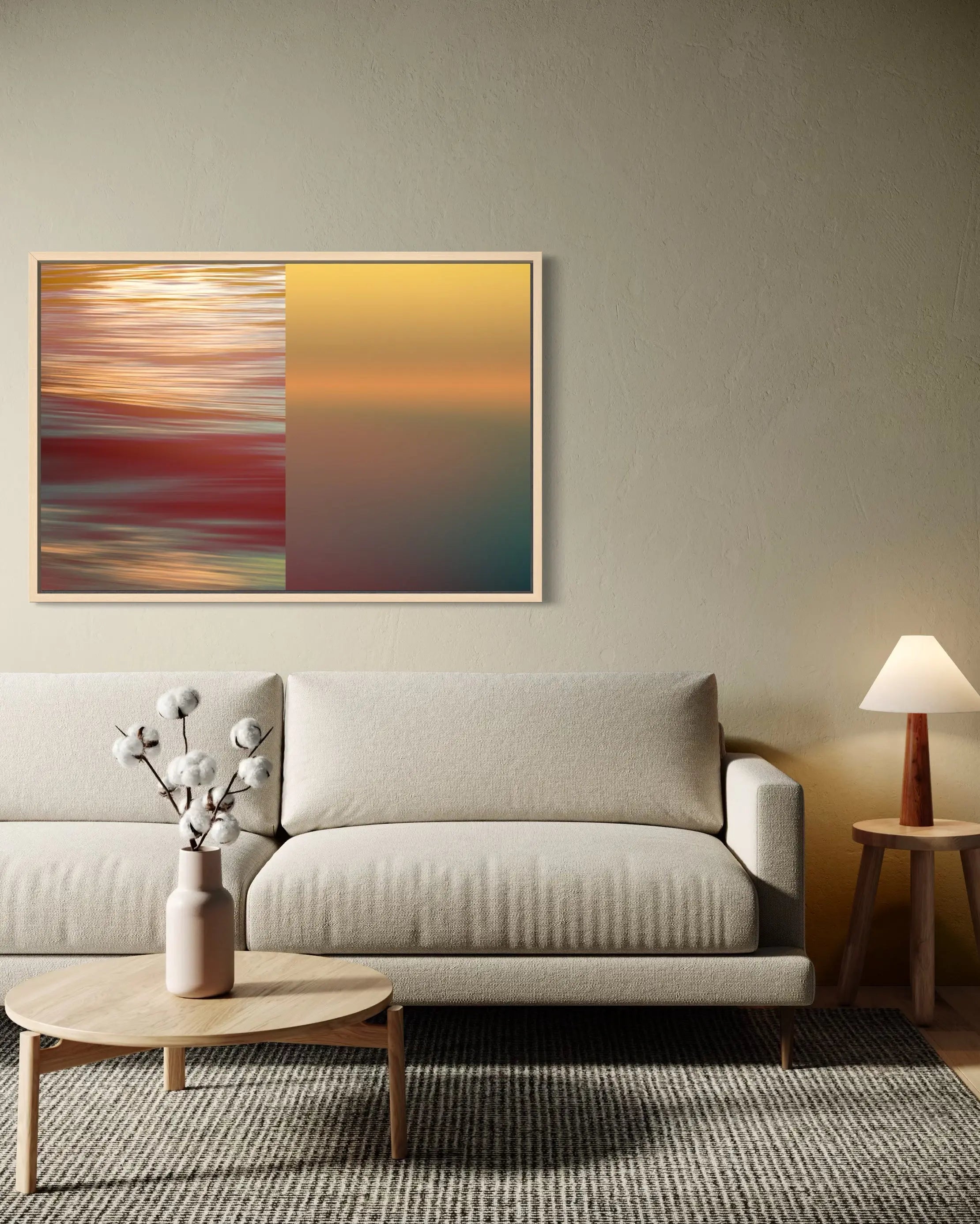 A framed abstract wall art print hangs above a light beige sofa in a styled living room. The artwork is split vertically into two sections: the left side features a blurred red, orange, and teal horizontal pattern, while the right side shows a smooth warm yellow-to-teal gradient.