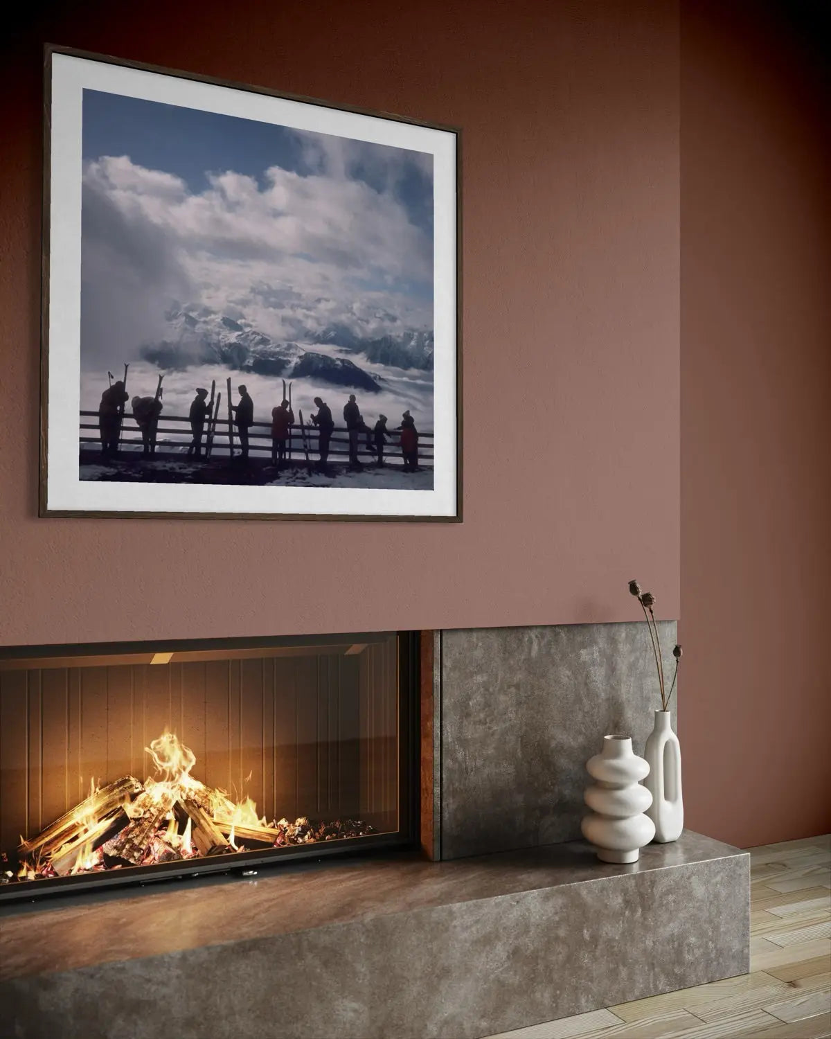 A framed landscape photograph of silhouetted figures against snowy mountains hangs on a terracotta-colored wall above a modern fireplace with a visible fire and stone surround.