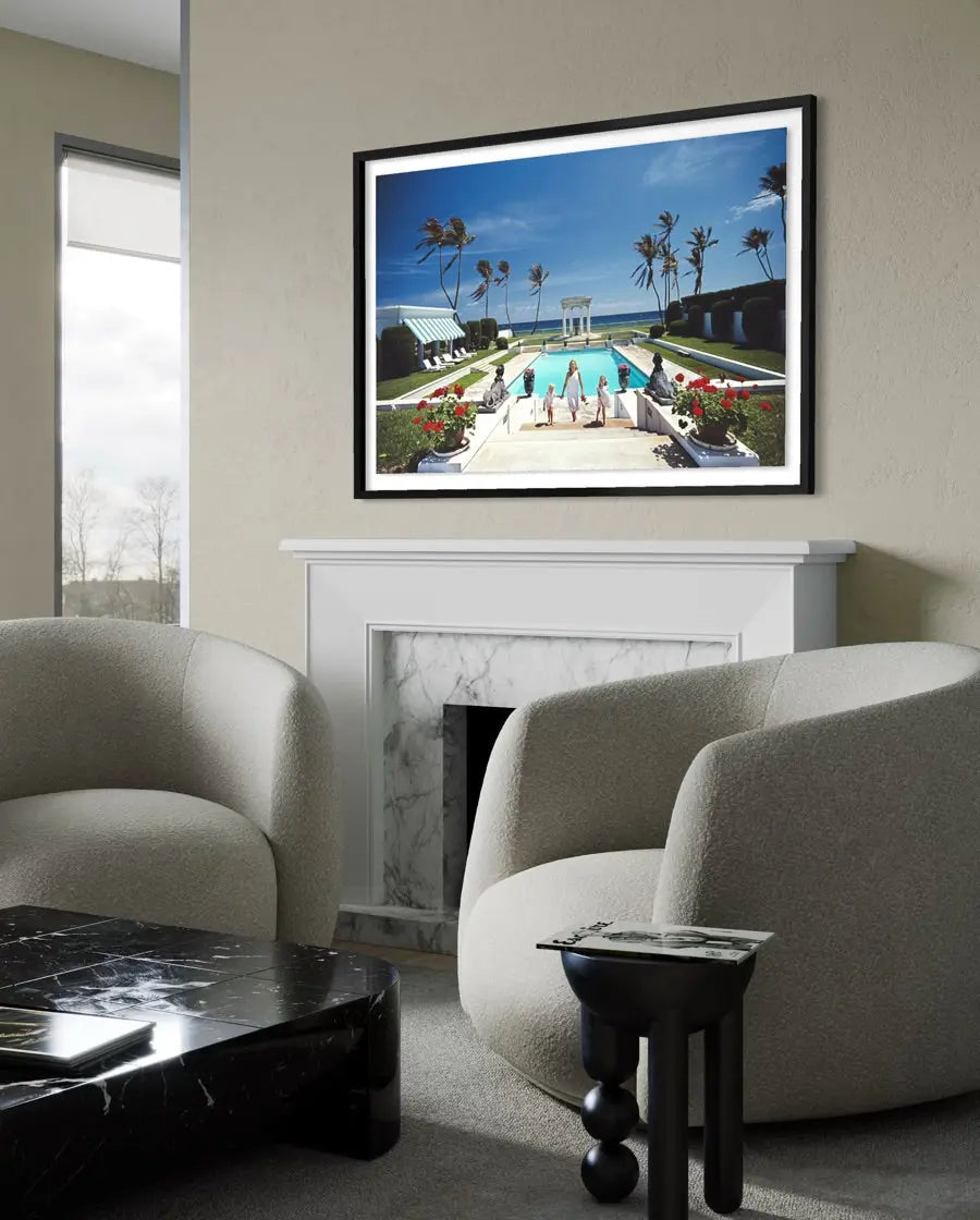 A framed art print depicting a luxurious poolside scene with palm trees and lounge chairs, displayed on a beige wall above a white fireplace in a modern living room setting. The Picturalist Slim Aarons Collection.