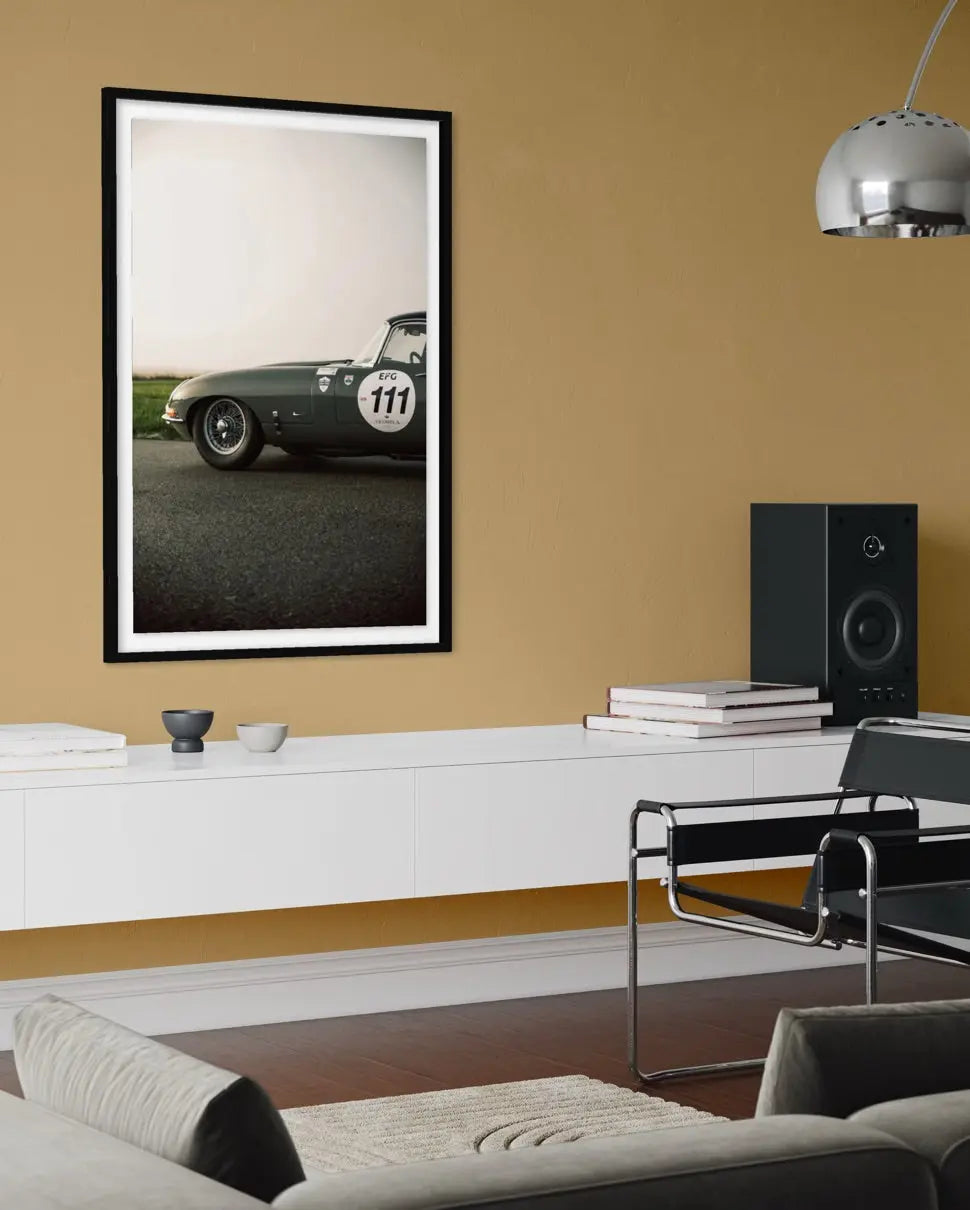 A framed vertical art print of a vintage green race car with the number 111, displayed on a warm mustard-yellow wall in a modern living room setting.