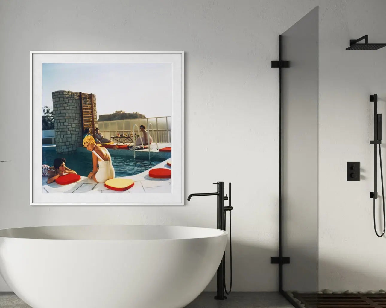 A framed art print depicting a vintage-style scene of a woman by a swimming pool with red float rings, displayed on a light gray bathroom wall above a white freestanding bathtub. The Picturalist Wall Art and Photography for Designed Interiors. Slim Aarons Collection, Poolside. Getty Images Gallery.