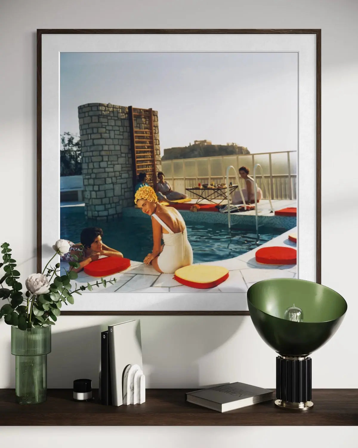 A framed art print depicting a vintage scene of people relaxing by a pool with red and yellow cushions, displayed on a shelf above a green bowl lamp and other decorative items.