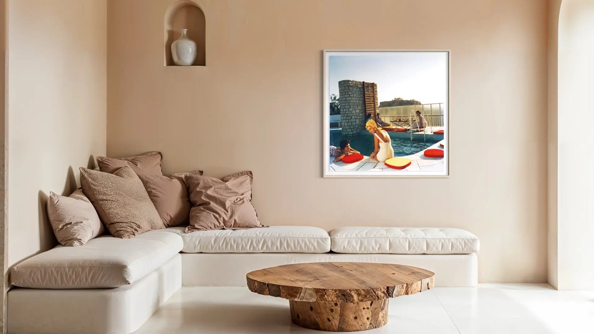 A framed art print depicting a woman in a yellow swimsuit by a pool, hanging on a beige wall above a modern white sectional sofa and a round wooden coffee table. The Picturalist Wall Art and Photography for Designed Interiors. Slim Aarons Collection, Poolside. Getty Images Gallery.