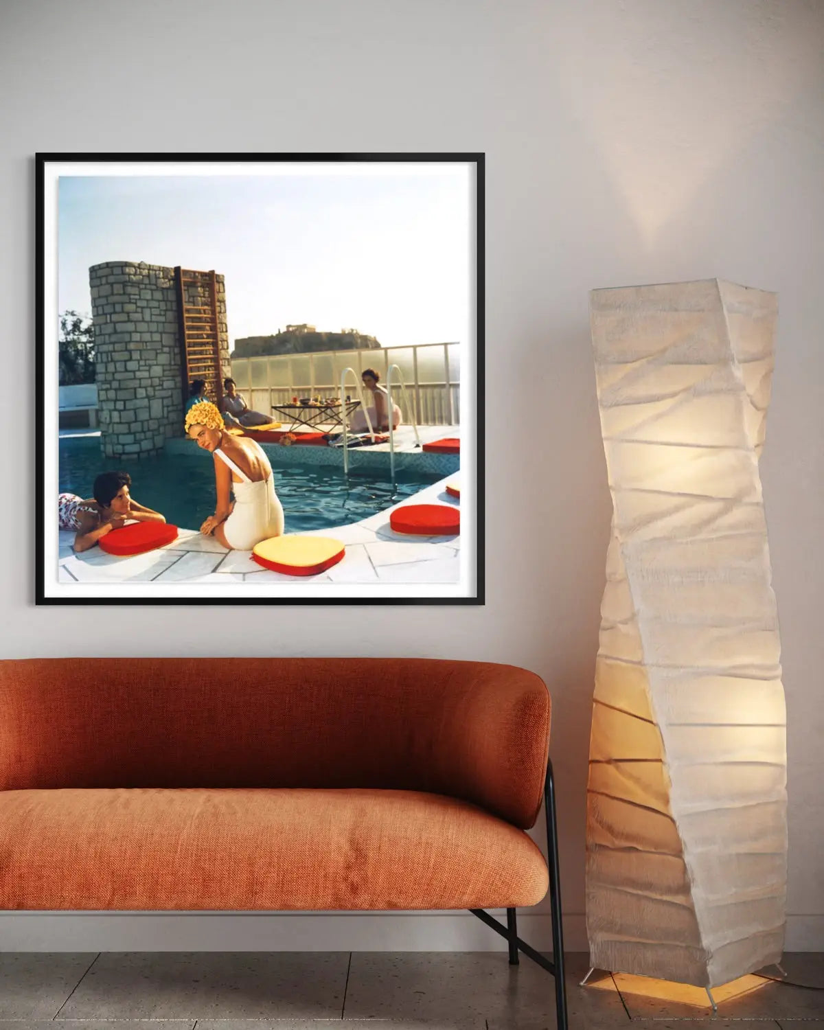 A framed art print depicting a retro scene of women by a swimming pool with red float rings, displayed on a wall above an orange-brown velvet sofa next to a tall floor lamp.