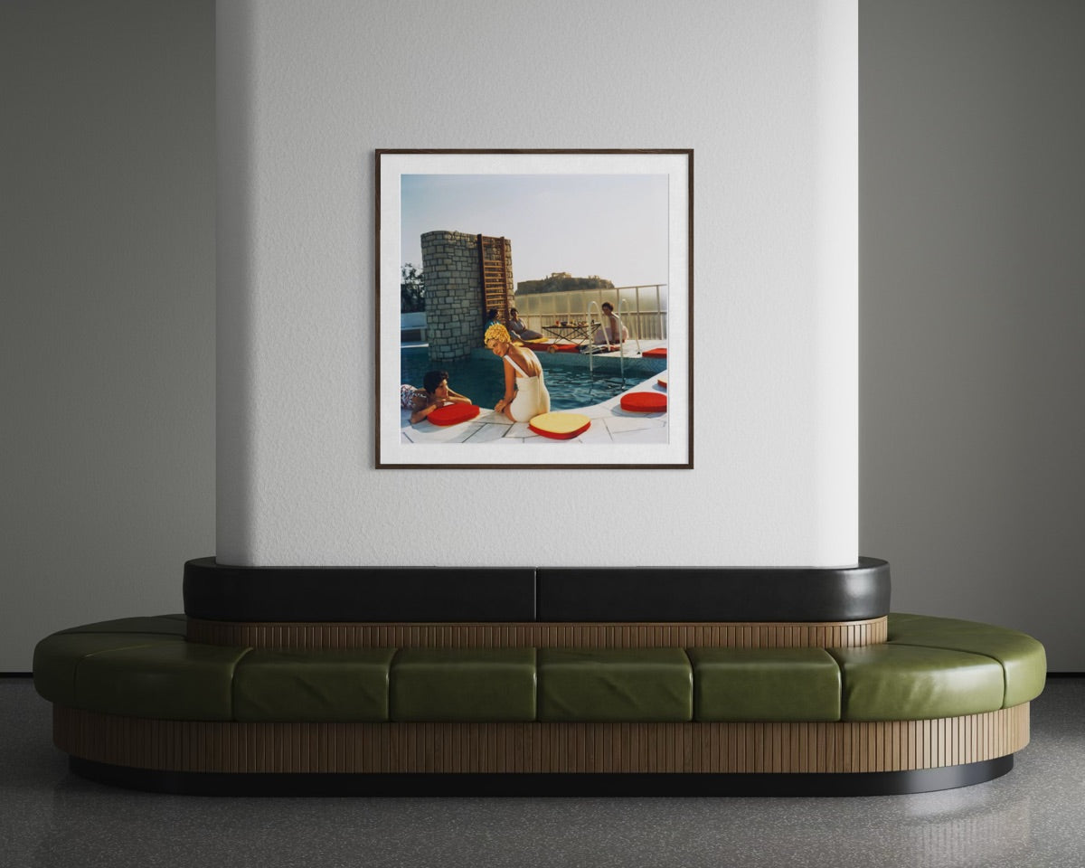 A framed art print depicting a retro scene of a woman by a pool with red bean bags, displayed on a white wall above a modern curved green and wood bench in a gray room.