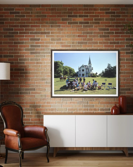 Framed picture of a group of people on a grassy field with a building in the background, displayed on a brick wall above a white cabinet.