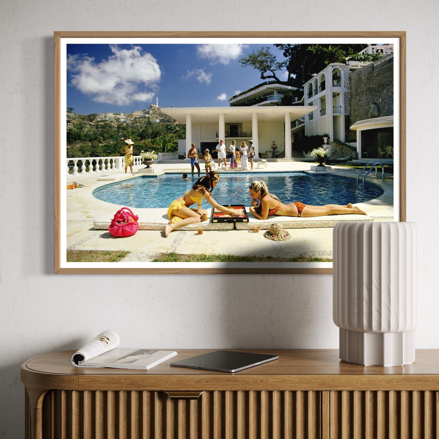 Framed artwork of people by a pool on a wall above a wooden console table.