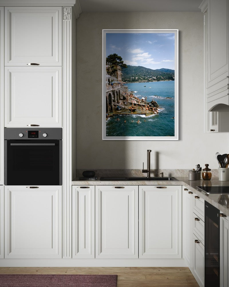 Modern kitchen with white cabinets, black appliances, and a large Slim Aarons photograph of Rapallo, Italy of a coastal scene.