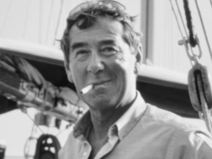 Black and white photo of a man with a pipe, wearing goggles on his head, standing in front of a boat.