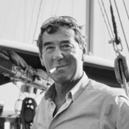 Black and white photo of a man with a pipe, wearing goggles on his head, standing in front of a boat.