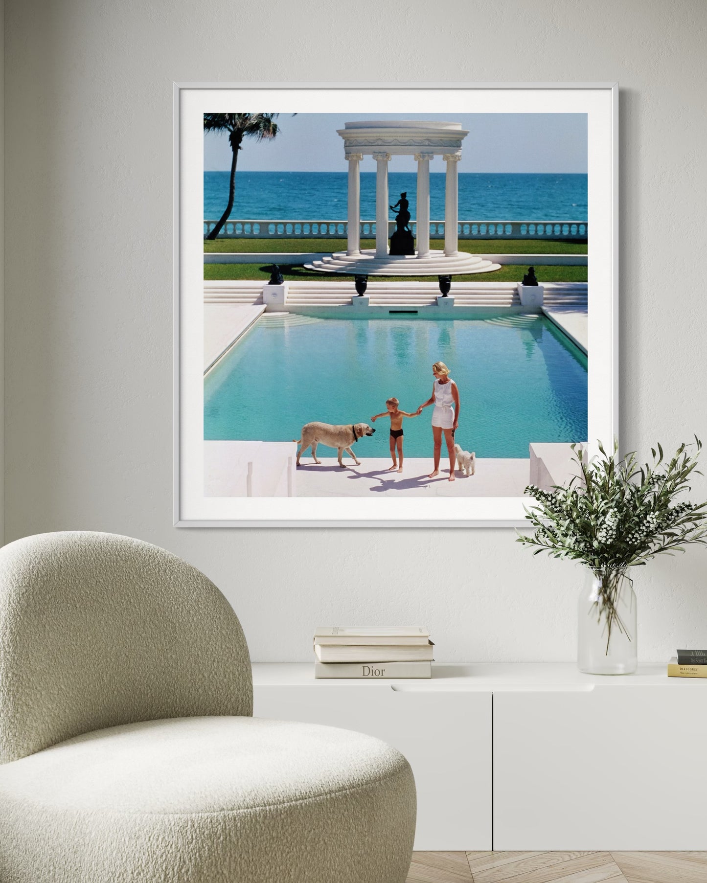 Framed photograph of a pool scene with people and dogs, displayed on a wall above a white cabinet.