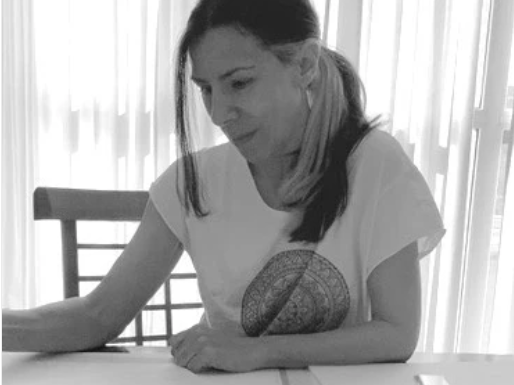 Woman sitting at a table with a decorative pillow on her lap, in a room with white curtains.