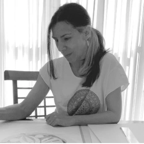 Woman sitting at a table with a decorative pillow on her lap, in a room with white curtains.