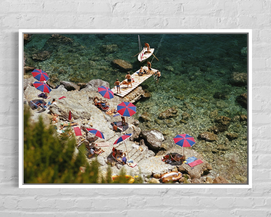 People sunbathing on rocks with colorful umbrellas near a body of water.