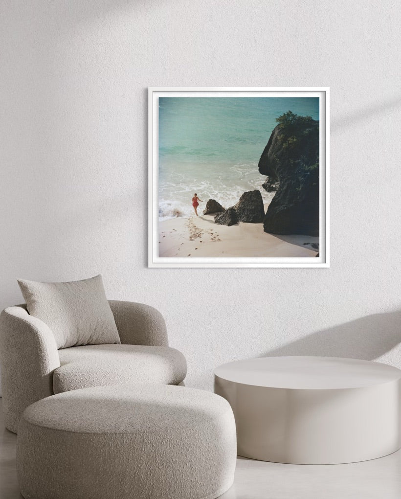 A framed square art print depicting a beach scene with turquoise water, dark rocks, and pale sand, displayed on a light wall above a beige armchair and round ottoman.