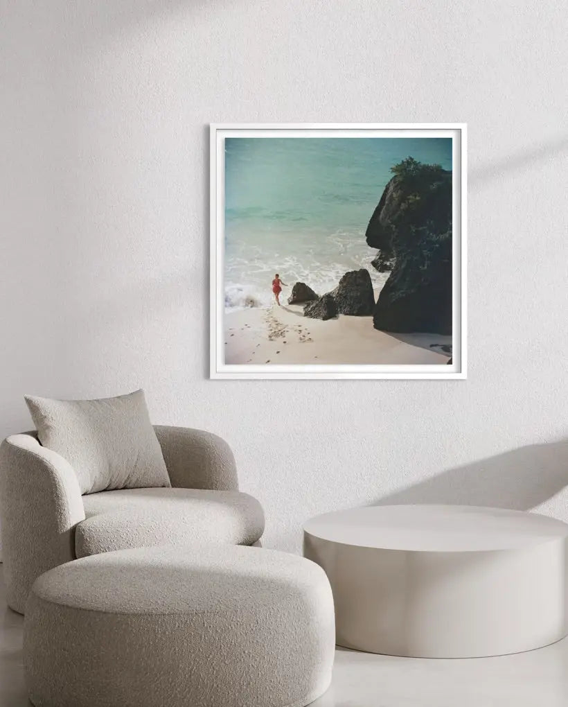 A framed square art print depicting a beach scene with turquoise water, dark rocks, and pale sand, displayed on a light wall above a beige armchair and round ottoman.