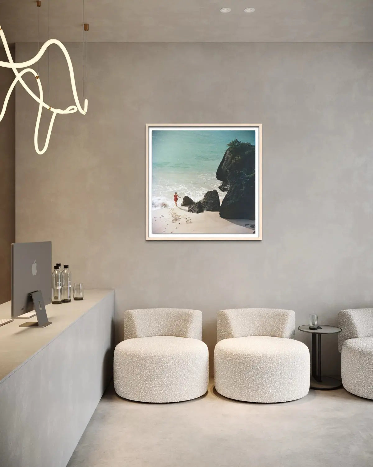 A framed square art print depicting a beach scene with turquoise water, white sand, and dark rocks, displayed on a textured gray wall in a modern minimalist interior with cream boucle chairs and a concrete counter.