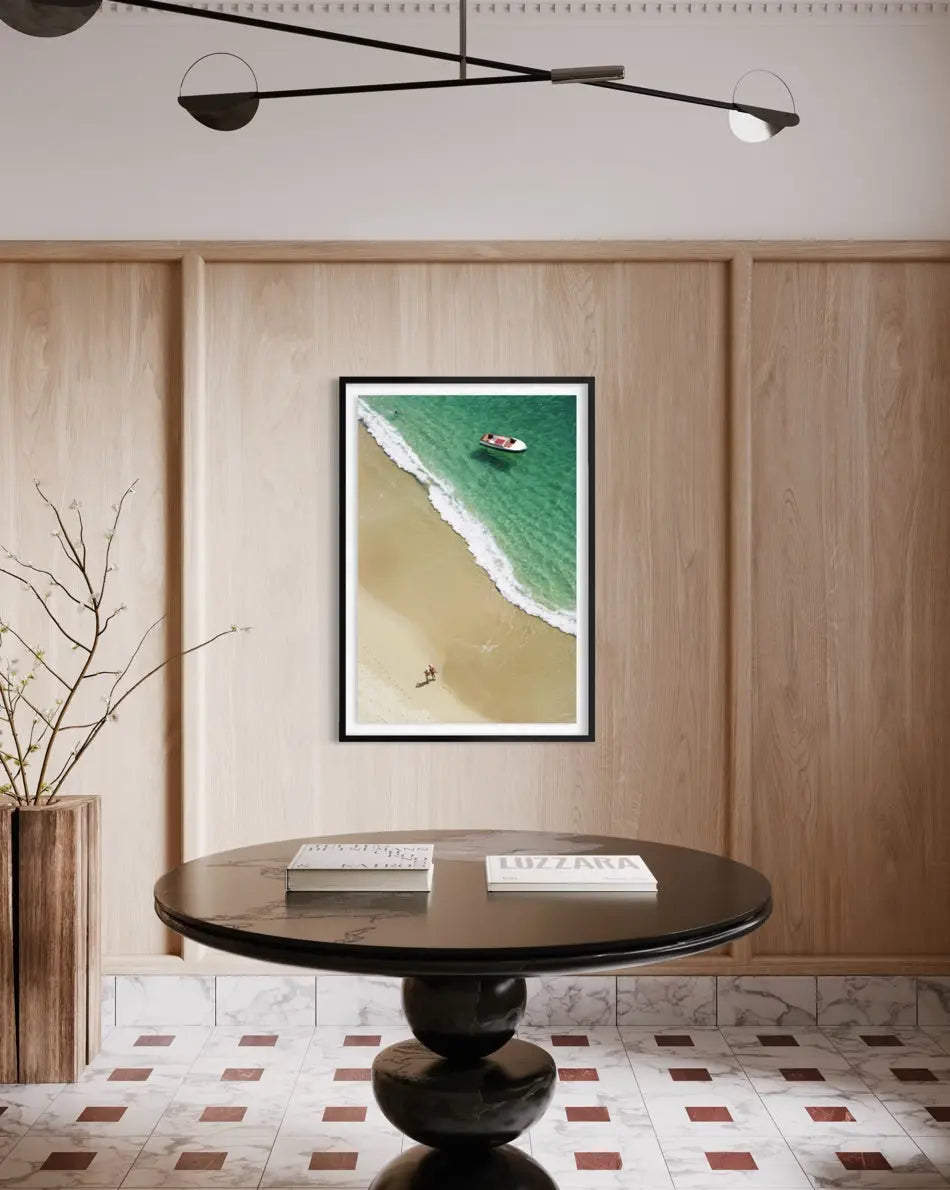 A framed vertical art print depicting an aerial view of a beach and turquoise ocean, displayed on a light wood paneled wall above a round black table in a modern interior setting.