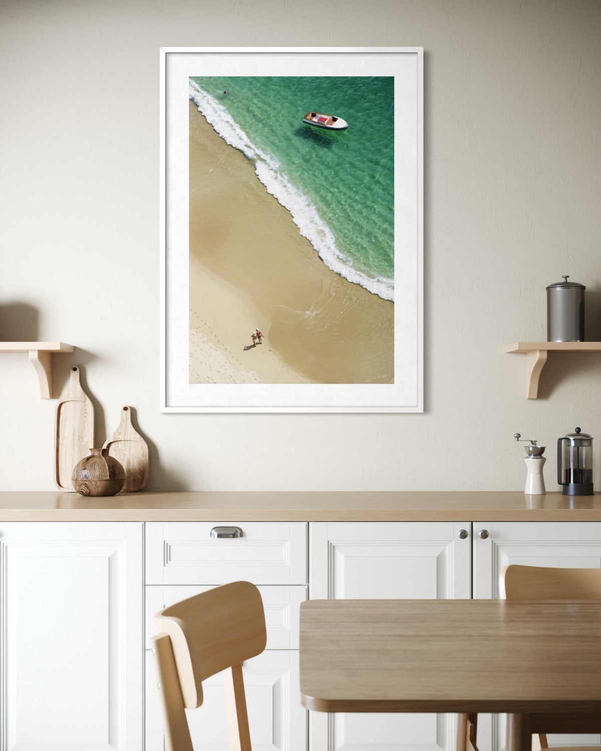 A framed vertical art print depicting an aerial view of a beach and turquoise ocean, displayed on a beige wall above a kitchen counter in a modern interior setting.