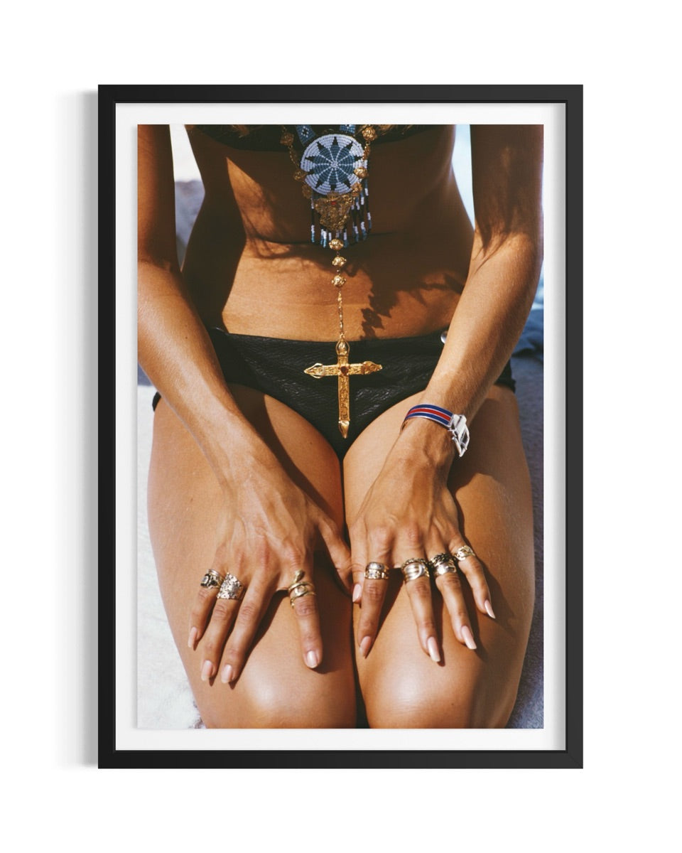 A framed art print featuring a close-up photograph of a woman wearing black bikini bottoms and multiple gold rings, with a black frame and white mat on a white background.