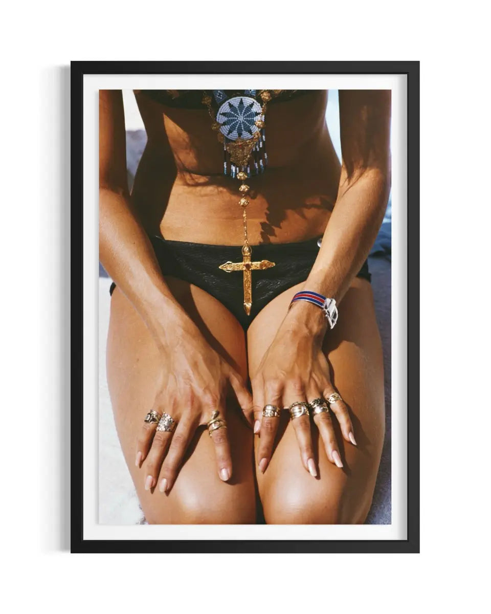 A framed art print featuring a close-up photograph of a woman wearing black bikini bottoms and multiple gold rings, with a black frame and white mat on a white background.