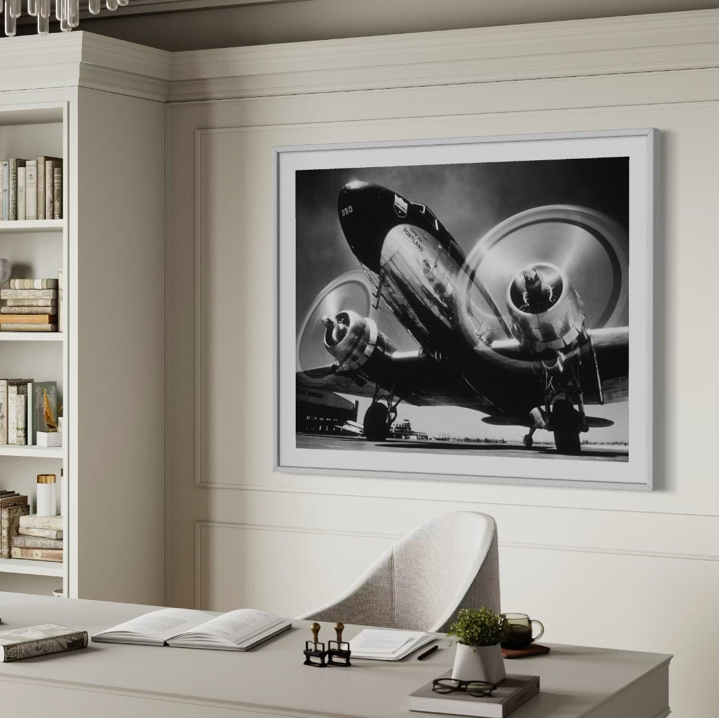 Framed black and white airplane photograph on a wall above a desk with books and decor.