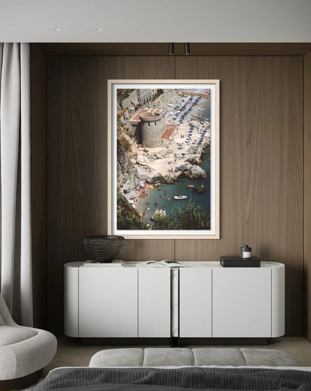 A framed vertical art print depicting a coastal seaside resort with a stone tower, blue pool water, and beach umbrellas, displayed on a dark wood wall above a white sideboard in a modern room.