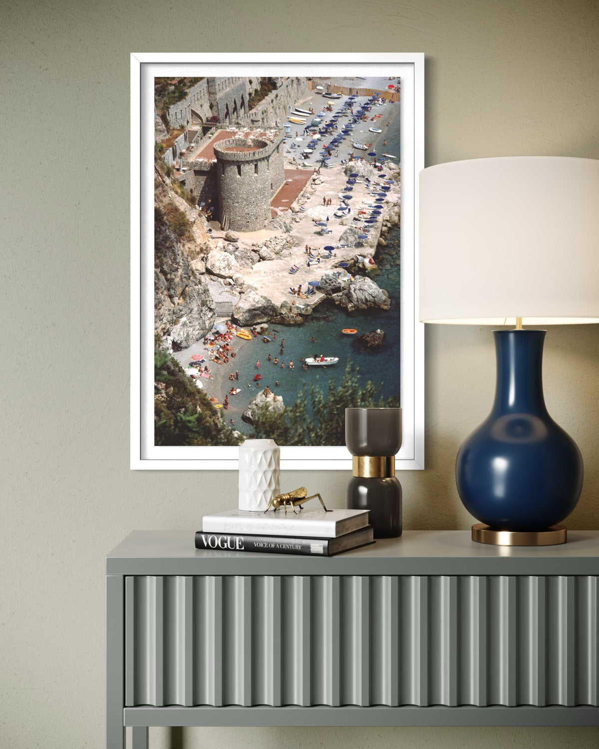 A framed vertical art print depicting an aerial view of a coastal beach and harbor with a stone tower, displayed on a gray console table next to a blue lamp and books.