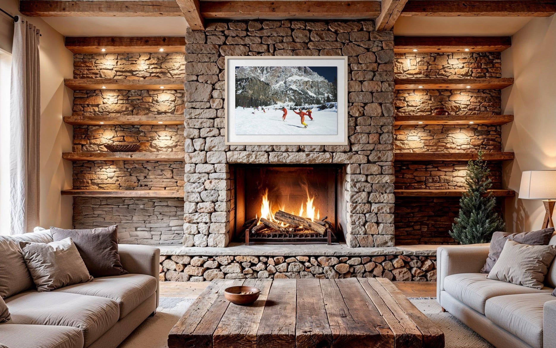 A cozy living room centered around a stone fireplace with a lit fire, wooden shelves lit from within, a rustic wood coffee table, two light beige sofas, and a framed winter mountain scene above the fireplace. The Picturalist Wall Art and Photography for Designed Interiors. Slim Aarons Collection, Skiing. Getty Images Gallery.