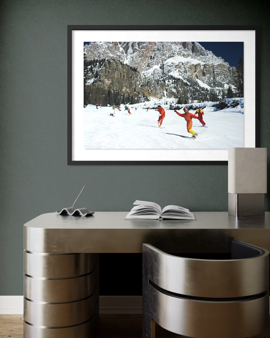 A framed art print depicting skiers in bright orange outfits on a snowy mountain slope, displayed on a dark gray wall above a modern metallic desk. The Picturalist Wall Art and Photography for Designed Interiors. Slim Aarons Collection, Skiing. Getty Images Gallery.