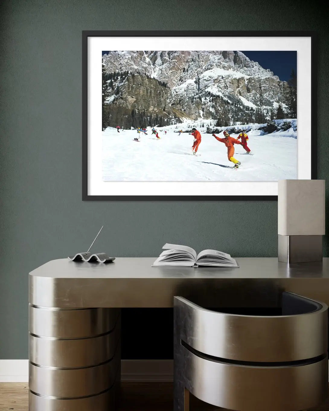 A framed art print depicting skiers in bright orange outfits on a snowy mountain slope, displayed on a dark gray wall above a modern metallic desk. The Picturalist Wall Art and Photography for Designed Interiors. Slim Aarons Collection, Skiing. Getty Images Gallery.