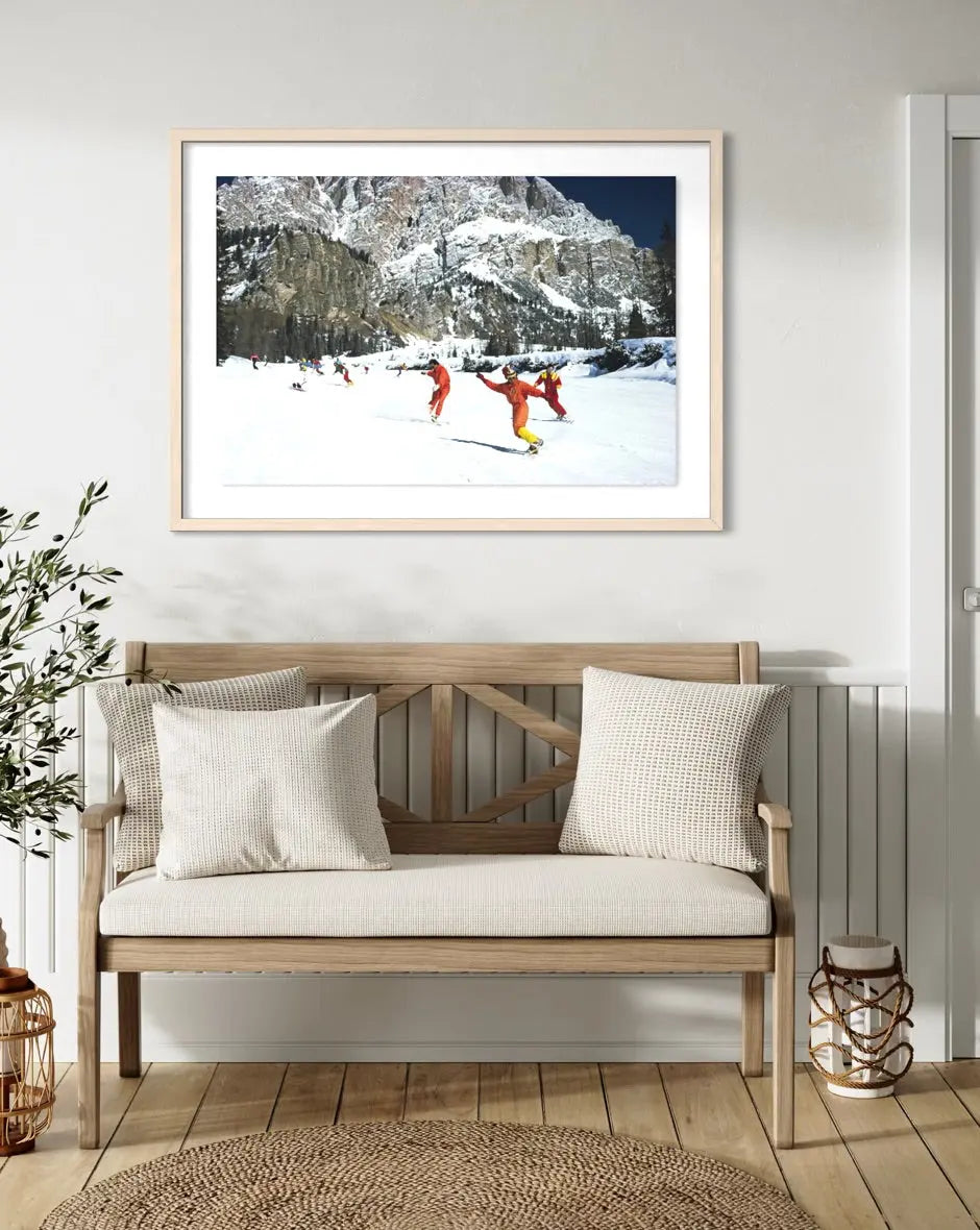 A framed art print depicting skiers in bright orange outfits against a snowy mountain landscape, displayed on a white wall above a wooden bench with neutral-toned cushions in a styled interior setting. The Picturalist Wall Art and Photography for Designed Interiors. Slim Aarons Collection, Skiing. Getty Images Gallery.
