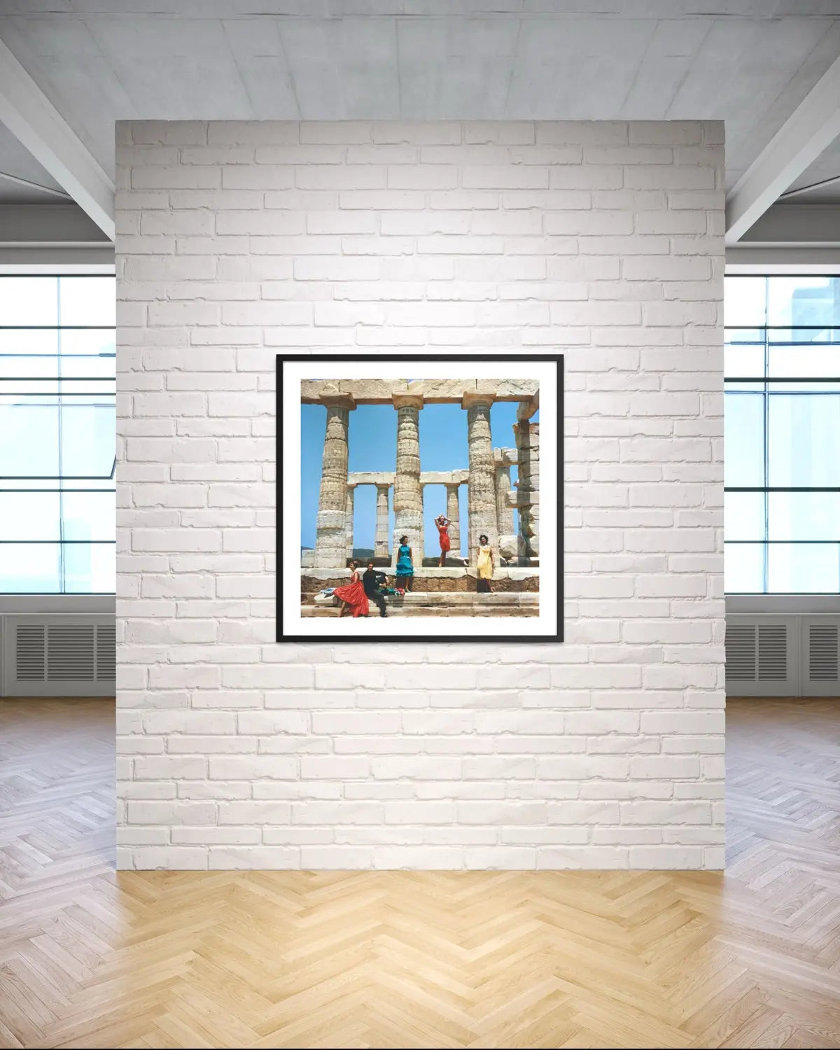 A framed art print depicting ancient stone columns and figures under a blue sky, displayed on a white brick wall in a modern room with wood floors.