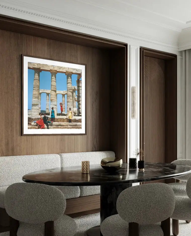 A framed art print depicting ancient stone columns and figures under a blue sky, displayed on a dark wood-paneled wall above a dining table with gray upholstered chairs.