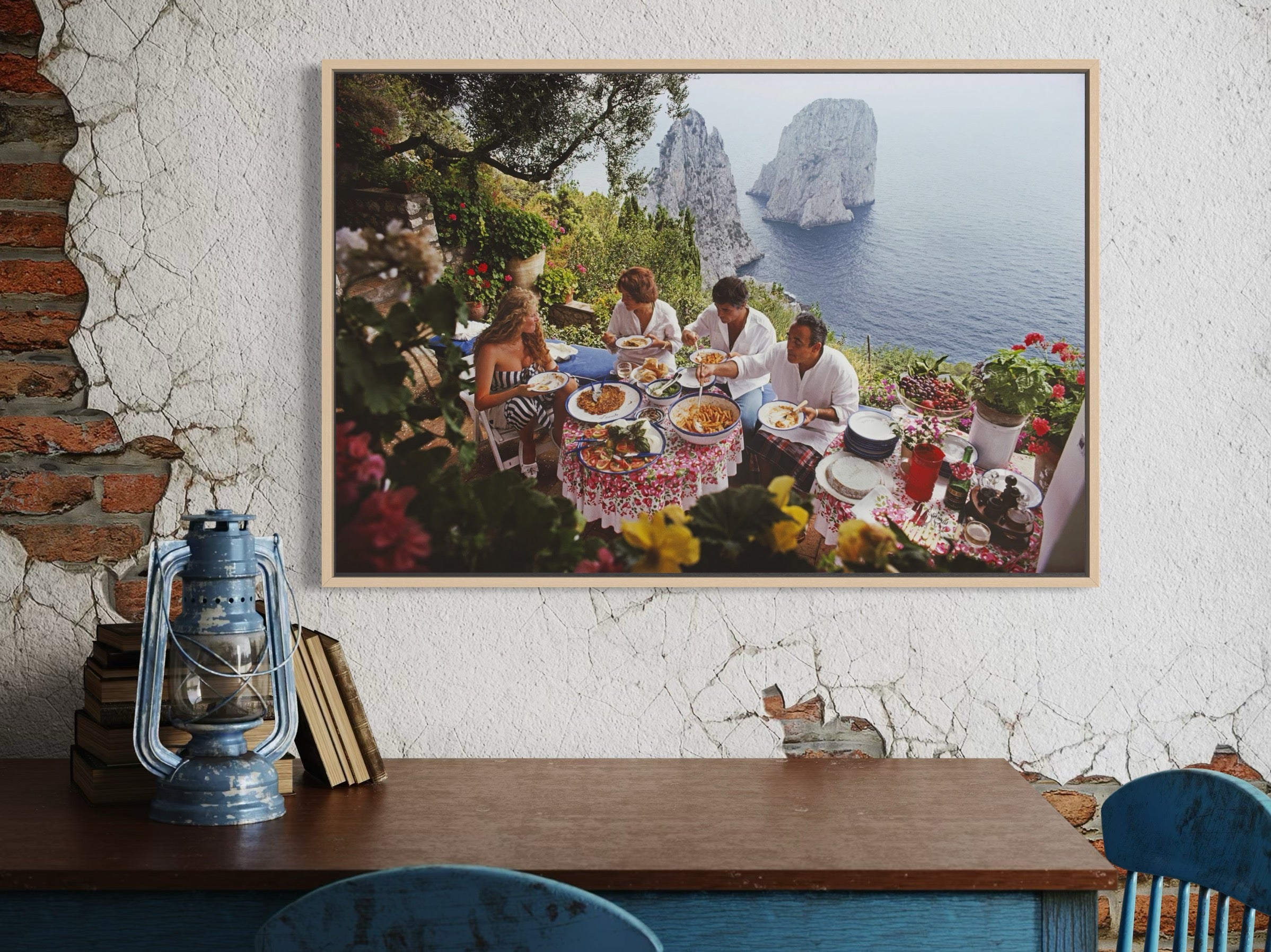 Framed artwork of a group dining outdoors with ocean view on a textured wall.