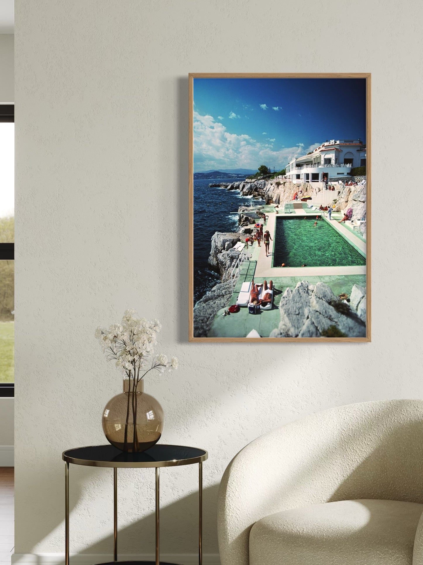 Framed photograph of a coastal scene with a pool on a wall above a side table and chair.