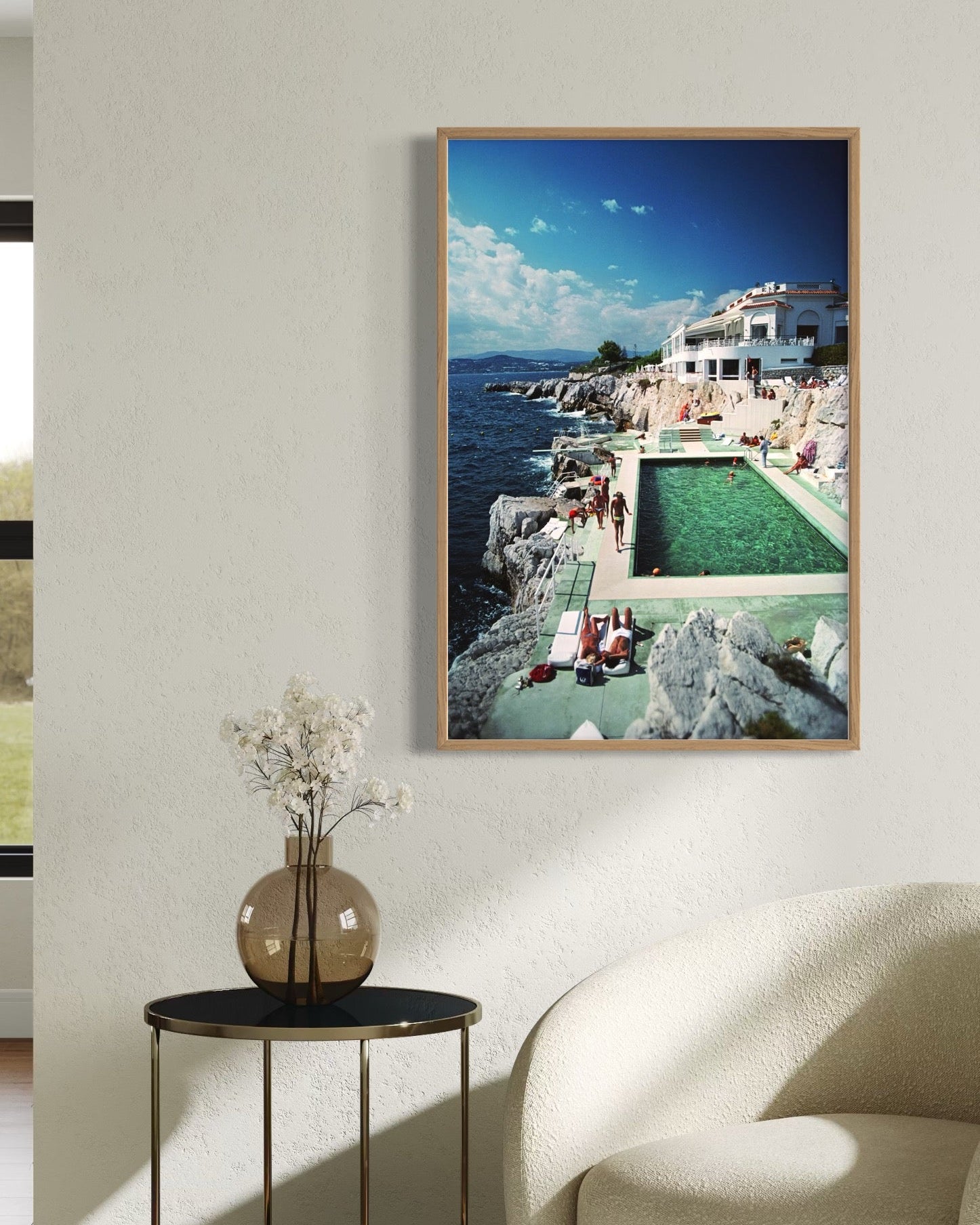 Framed photograph of a coastal scene with a pool on a wall above a side table and chair.