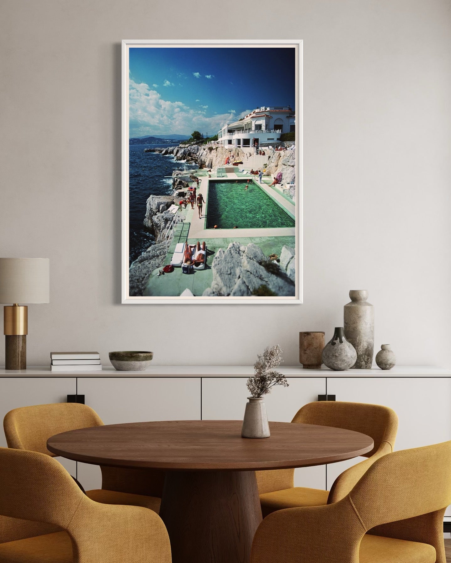 Dining room with a round wooden table and yellow chairs, featuring a framed picture of a poolside scene on the wall.