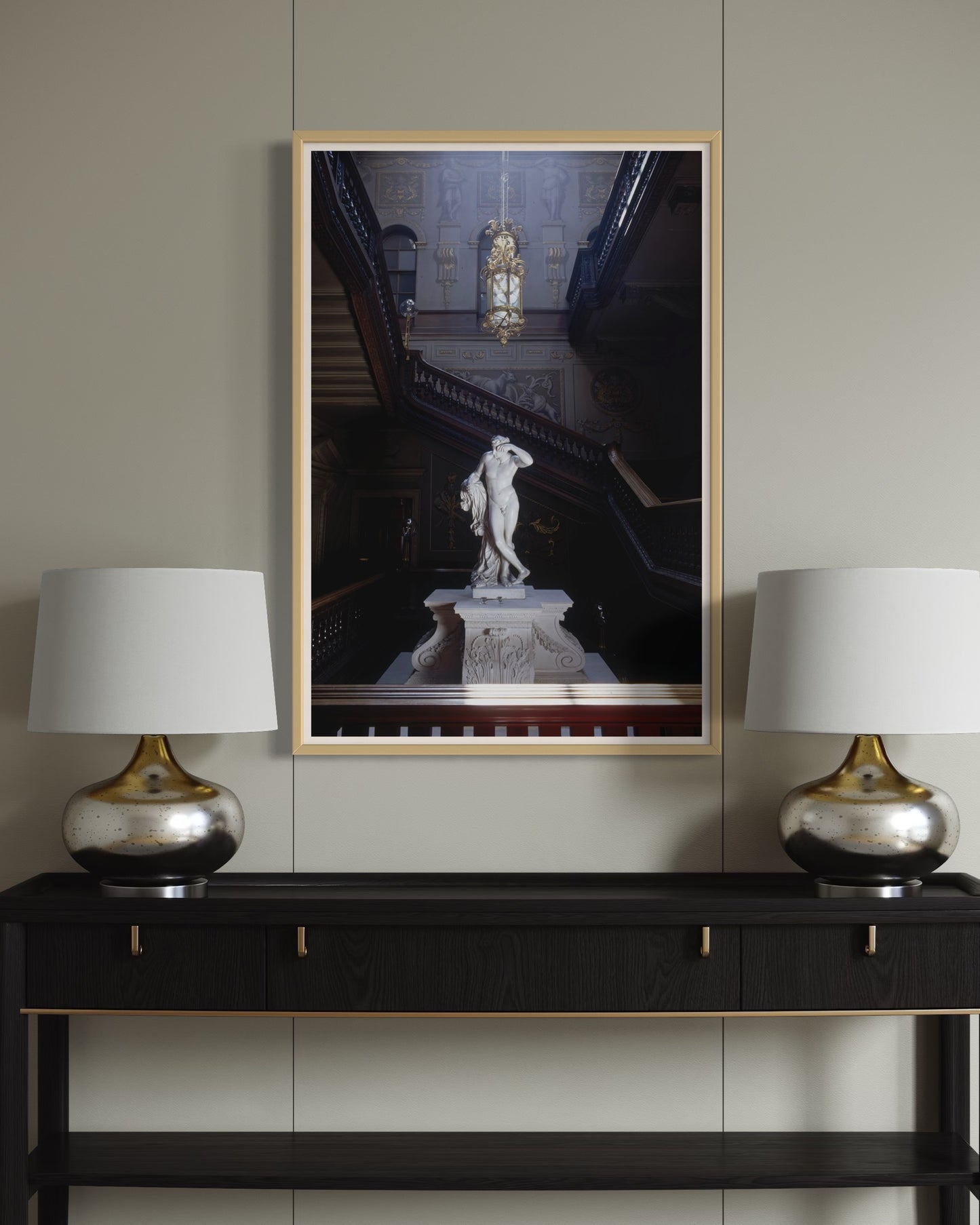 Framed artwork of a statue on a wall above a black console table with two silver lamps.