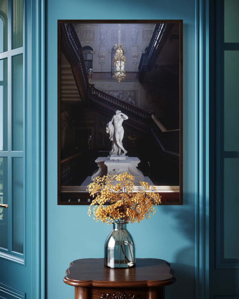Framed artwork of a classical statue on a blue wall with a vase of flowers below.