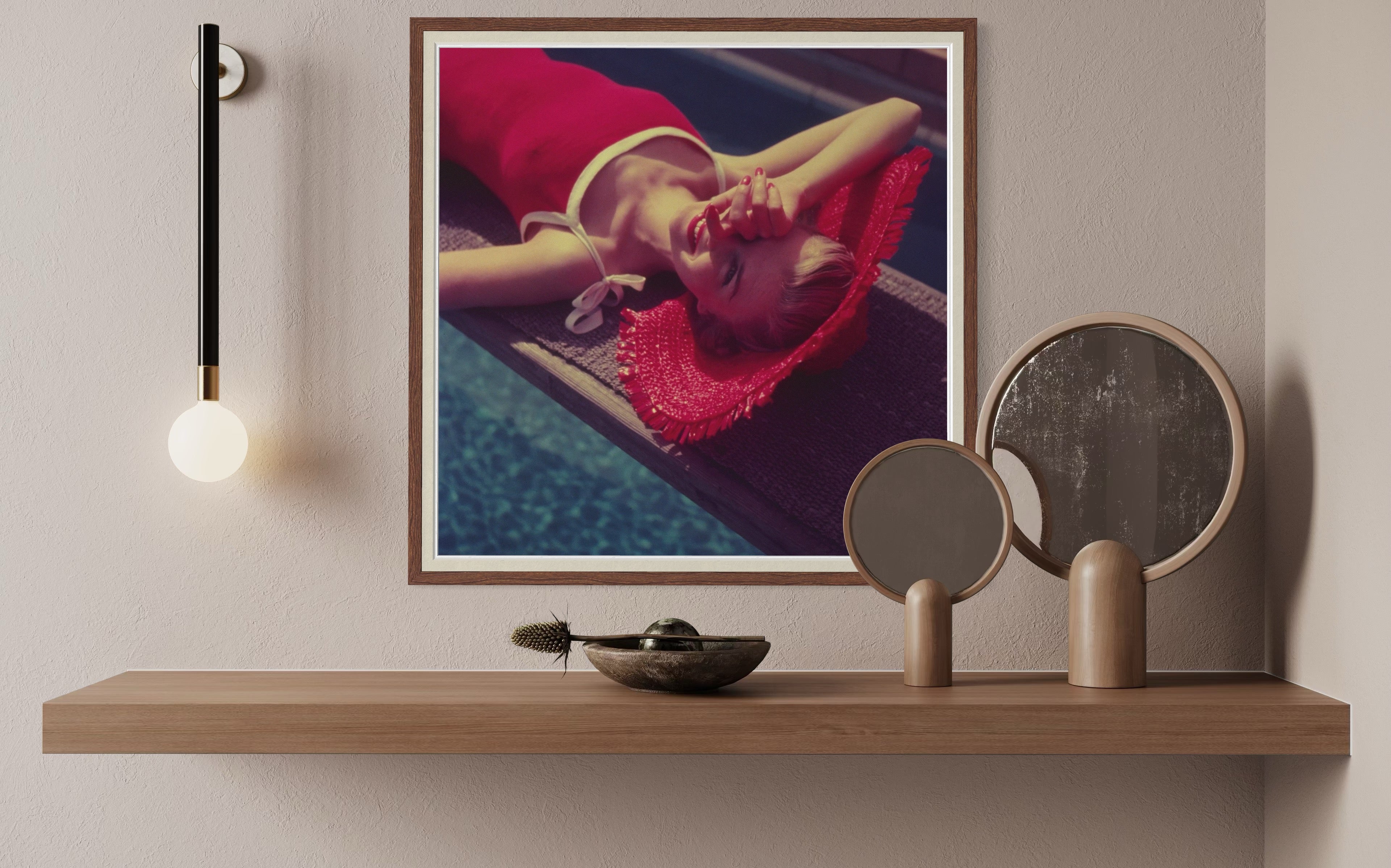 Framed artwork of a person in a red dress lying on a poolside, displayed on a wooden shelf with decorative items.